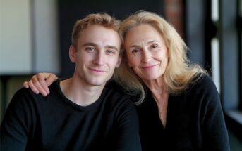 A smiling older woman with long blonde hair sits next to a younger man with short light brown hair. They both wear black sweaters and look at the camera, with the woman’s arm around the man’s shoulders.