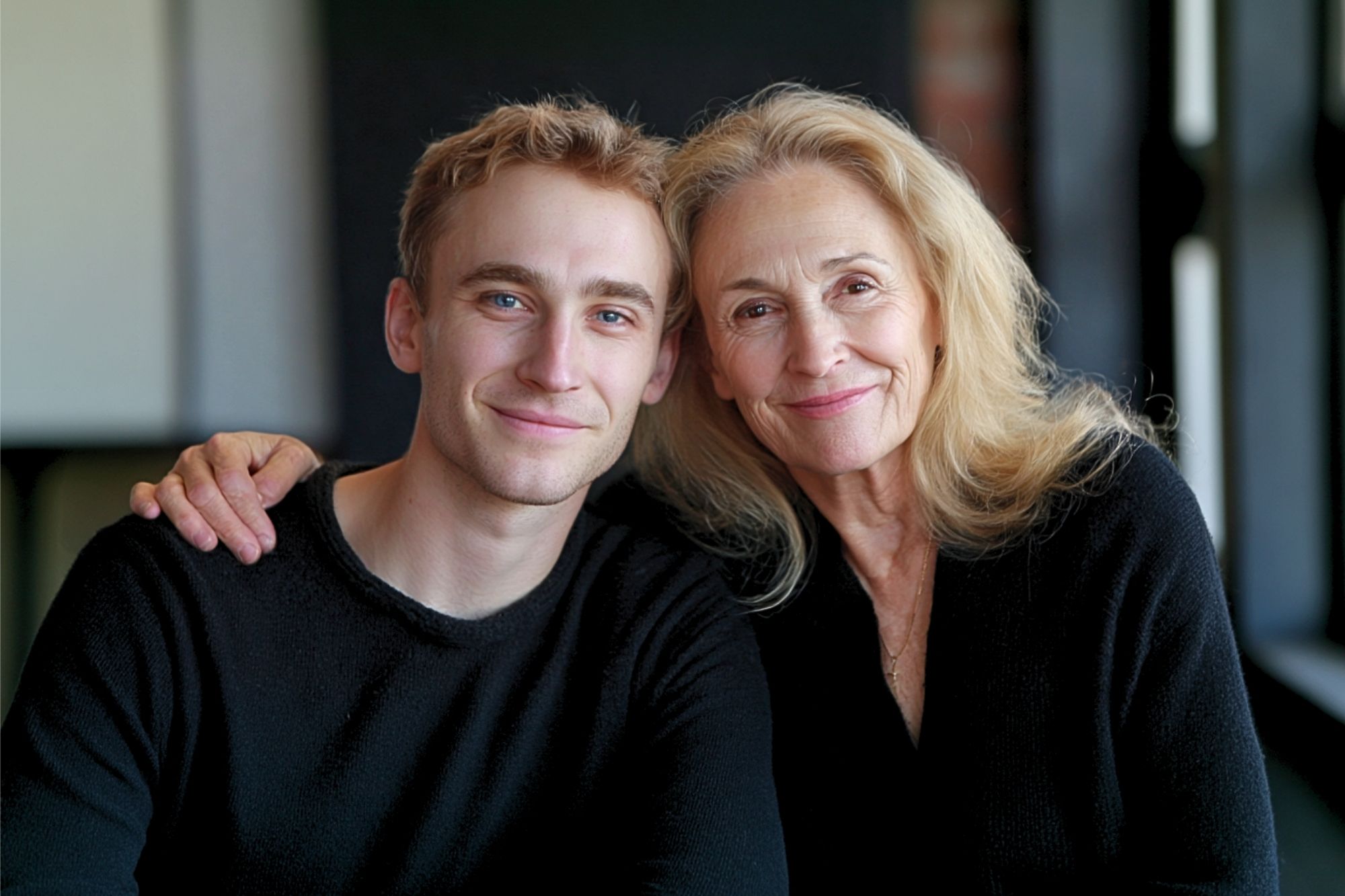 A smiling older woman with long blonde hair sits next to a younger man with short light brown hair. They both wear black sweaters and look at the camera, with the woman’s arm around the man’s shoulders.