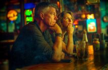 A man and woman sit at a dimly lit bar, both appearing thoughtful as they gaze ahead. Drinks and bottles are on the counter, with colorful neon signs glowing in the blurred background.