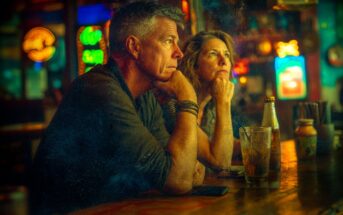 A man and woman sit at a dimly lit bar, both appearing thoughtful as they gaze ahead. Drinks and bottles are on the counter, with colorful neon signs glowing in the blurred background.