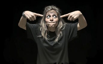 A woman with long, wavy hair in a black t-shirt points at her head with both index fingers and looks upward with a playful, puzzled expression against a black background.
