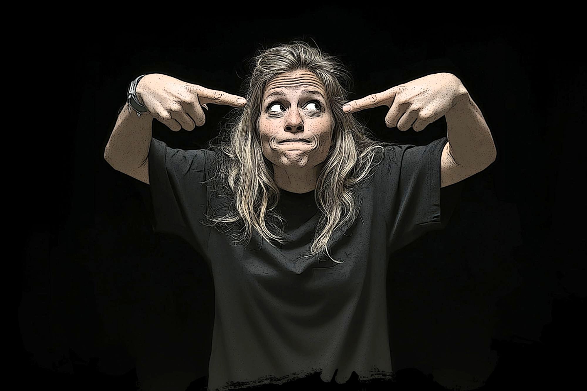 A woman with long, wavy hair in a black t-shirt points at her head with both index fingers and looks upward with a playful, puzzled expression against a black background.
