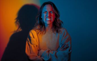 A woman with wavy hair crosses her arms and clenches her teeth in a tense expression. She is lit by dramatic orange and blue lighting, casting a shadow on the wall behind her.