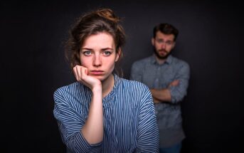 A woman with a sad expression rests her chin on her hand in the foreground, while a man with crossed arms stands in the blurred background against a dark backdrop.