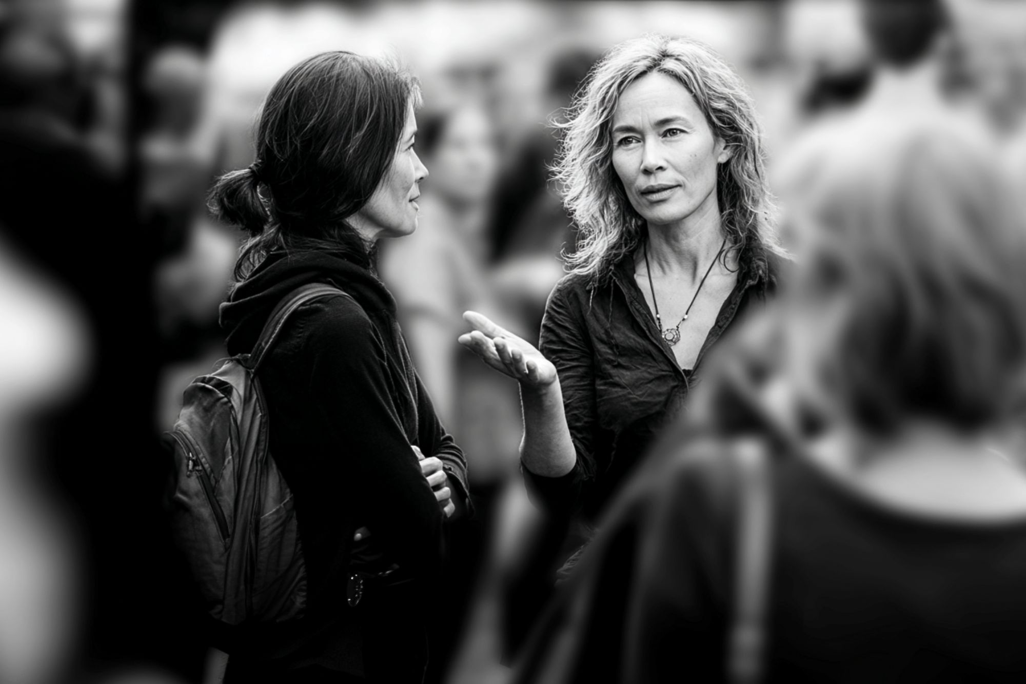 Two women stand facing each other in conversation outdoors, surrounded by a blurred crowd. One woman gestures with her hand while speaking; both are dressed casually. The image is in black and white.