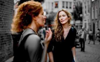 Two women talk on a cobblestone street; one is in sharp focus while the other is blurred in the foreground. The background is desaturated, while the women are in color.