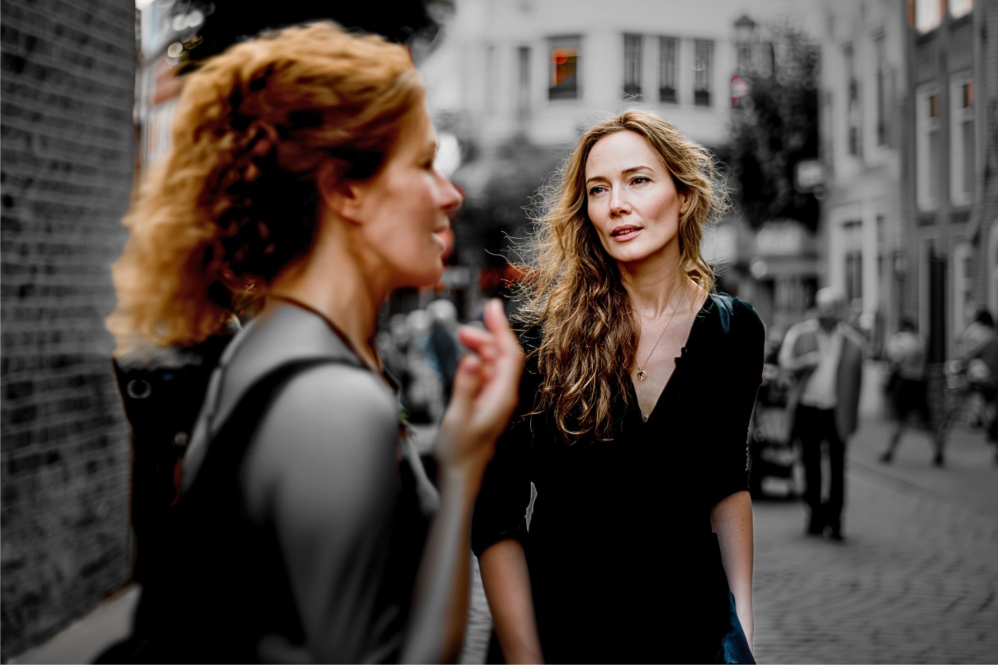 Two women talk on a cobblestone street; one is in sharp focus while the other is blurred in the foreground. The background is desaturated, while the women are in color.