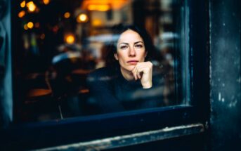 A woman sits inside a cafe, looking thoughtfully out a window with her chin resting on her hand. Warm, blurred lights and reflections fill the background, creating a cozy atmosphere.