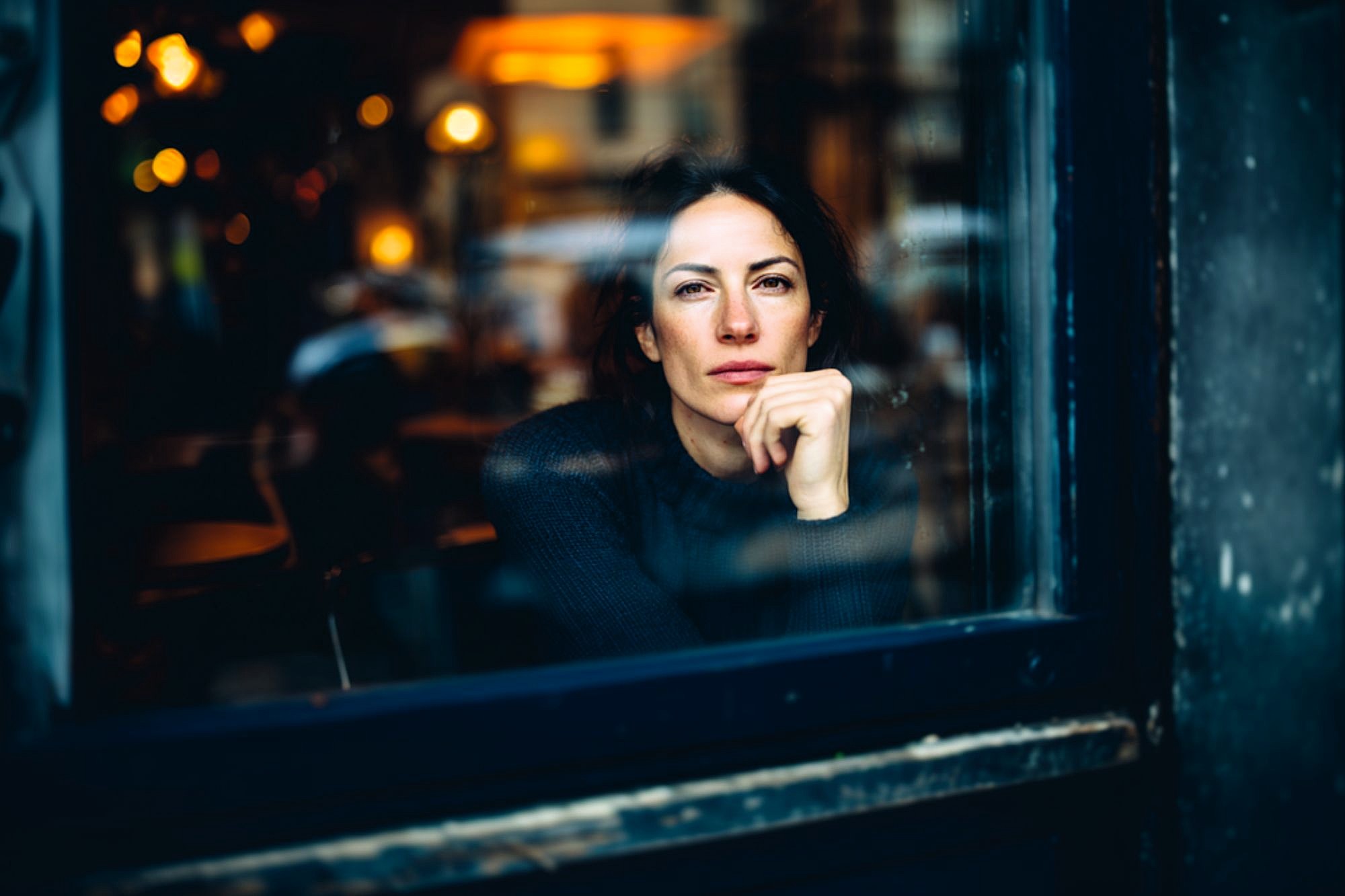 A woman sits inside a cafe, looking thoughtfully out a window with her chin resting on her hand. Warm, blurred lights and reflections fill the background, creating a cozy atmosphere.