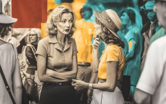 Two women in vintage clothing have a serious conversation outdoors, surrounded by people. The background features artistic, colorful painted elements in teal, orange, and red tones.