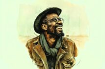 A smiling man with a beard, wearing glasses, a hat, a scarf, and a brown jacket, stands against a light, abstract background.