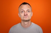 A man with short hair and a gray T-shirt looks up with a slightly concerned or uncertain expression, standing against an orange background.