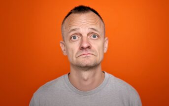 A man with short hair and a gray T-shirt looks up with a slightly concerned or uncertain expression, standing against an orange background.