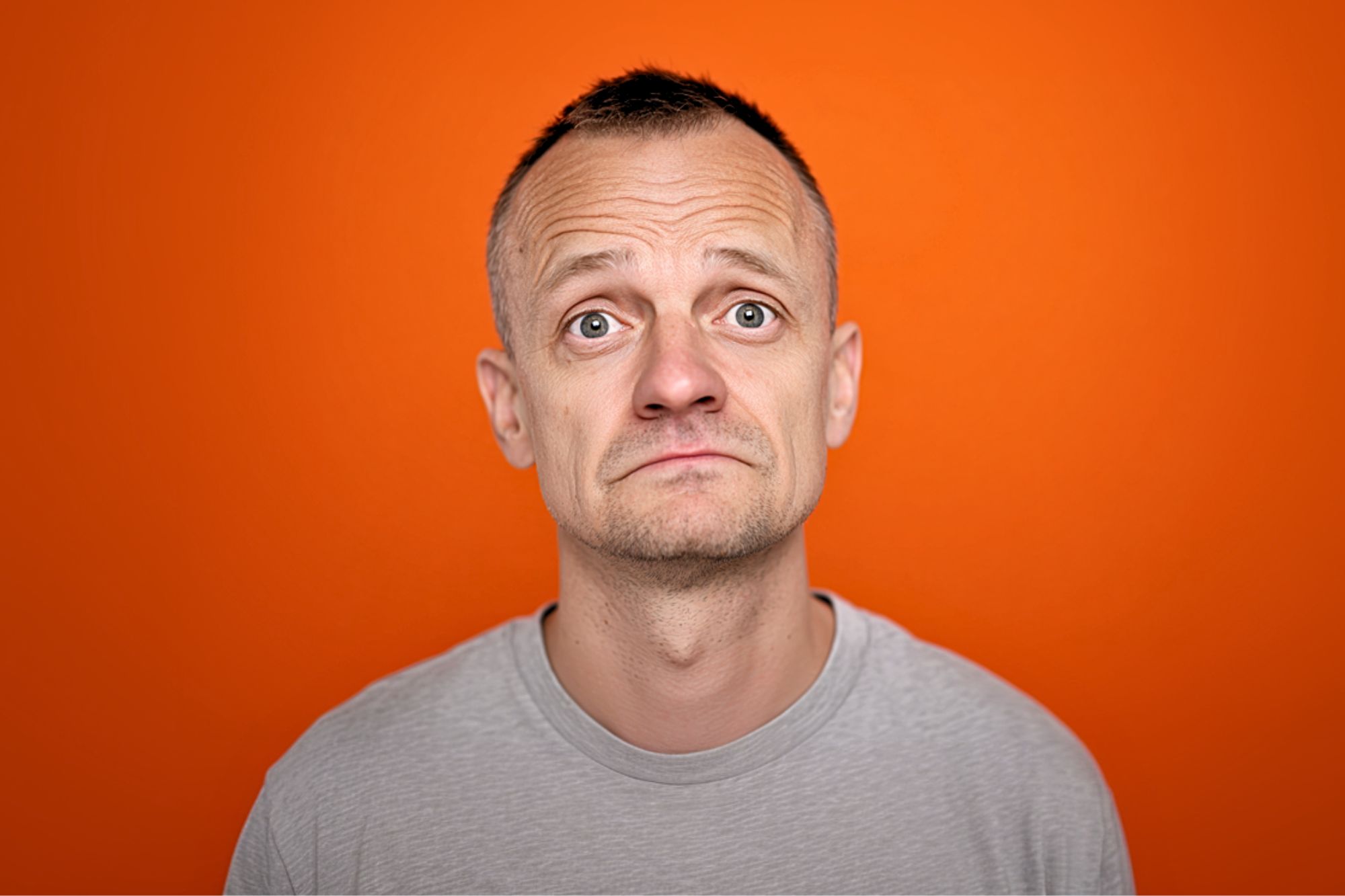 A man with short hair and a gray T-shirt looks up with a slightly concerned or uncertain expression, standing against an orange background.