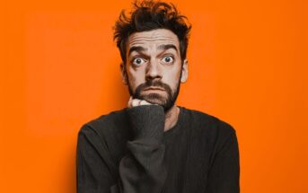 A man with messy hair and a beard, wearing a dark shirt, rests his chin on his hand while staring wide-eyed at the camera against a bright orange background.