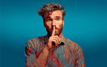 A man with tousled hair and a beard, wearing a colorful striped shirt, holds his index finger to his lips in a “shh” gesture against a blue background. The image has a digital, stylized effect.