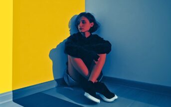A woman with short hair sits on the floor in a corner, hugging her knees. Blue and yellow lighting divides the walls and floor, casting dramatic shadows and creating a moody, artistic atmosphere.