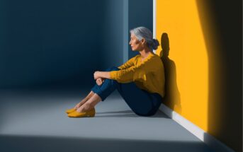 A woman with gray hair sits on the floor against a yellow wall, hugging her knees and looking thoughtful. She wears a yellow blouse, blue pants, and yellow shoes. Sunlight casts her shadow on the wall.