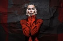 A woman with messy hair wearing a bright red turtleneck holds her face in her hands and looks up with a distressed, worried expression. The crumpled dark background emphasizes her dramatic pose.