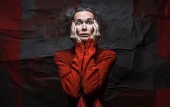 A woman with messy hair wearing a bright red turtleneck holds her face in her hands and looks up with a distressed, worried expression. The crumpled dark background emphasizes her dramatic pose.