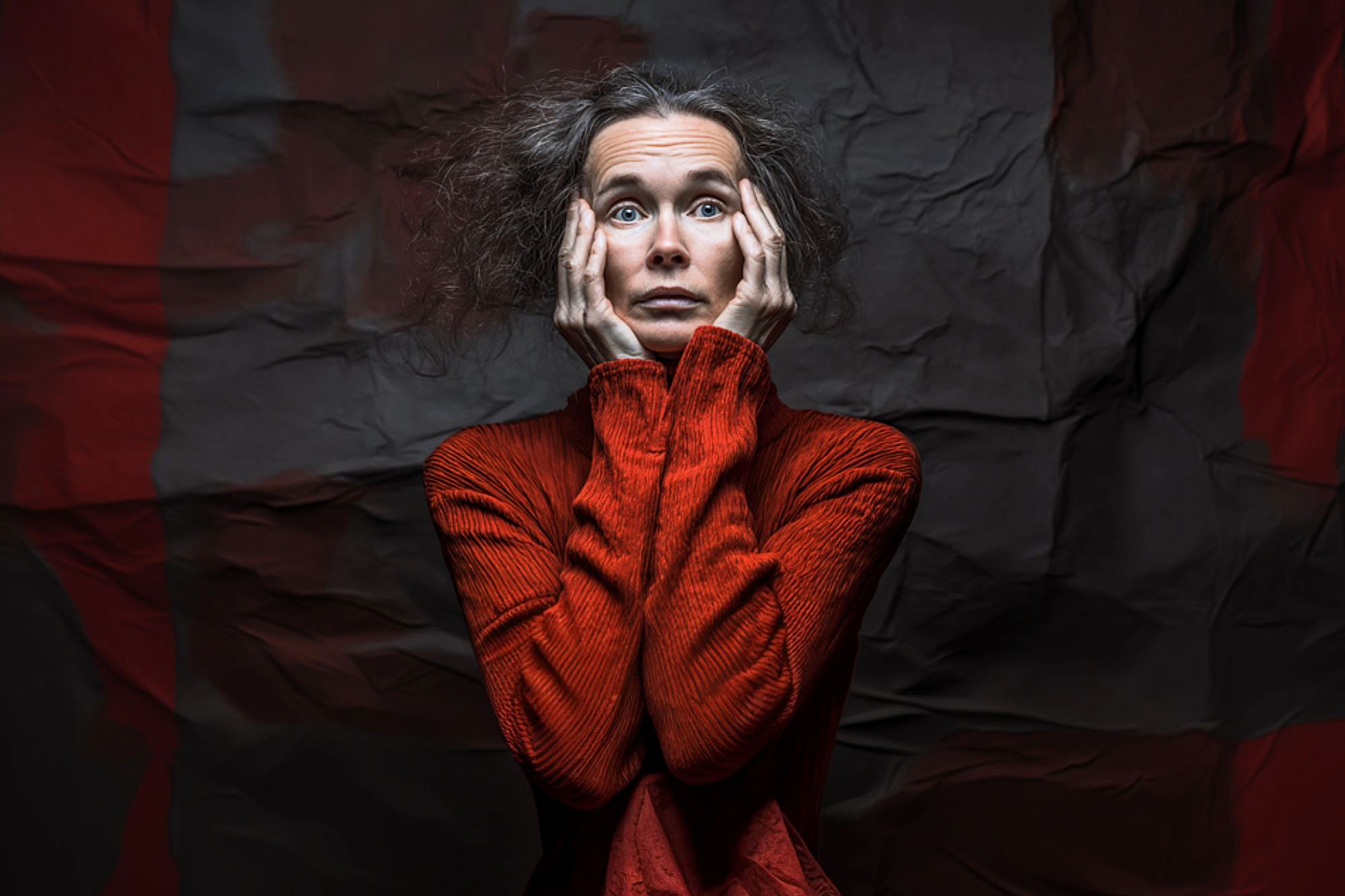 A woman with messy hair wearing a bright red turtleneck holds her face in her hands and looks up with a distressed, worried expression. The crumpled dark background emphasizes her dramatic pose.