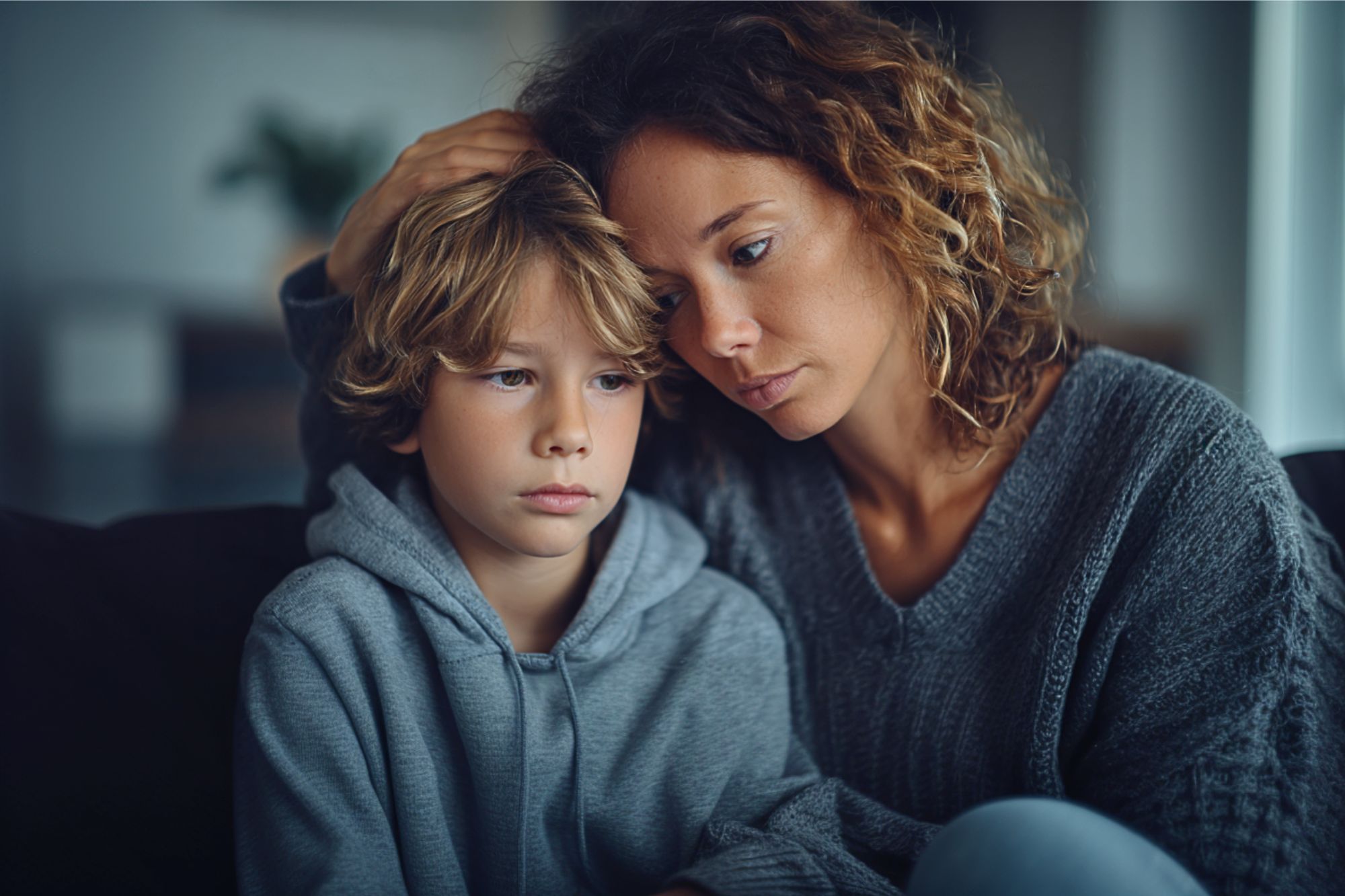 A woman sits closely with a young boy, gently resting her hand on his head. Both appear thoughtful and concerned, sitting together on a couch in a softly lit room.
