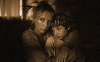 A woman hugs a young boy closely, both looking somber and thoughtful. The warm, sepia-toned lighting and soft focus create an intimate, comforting atmosphere.