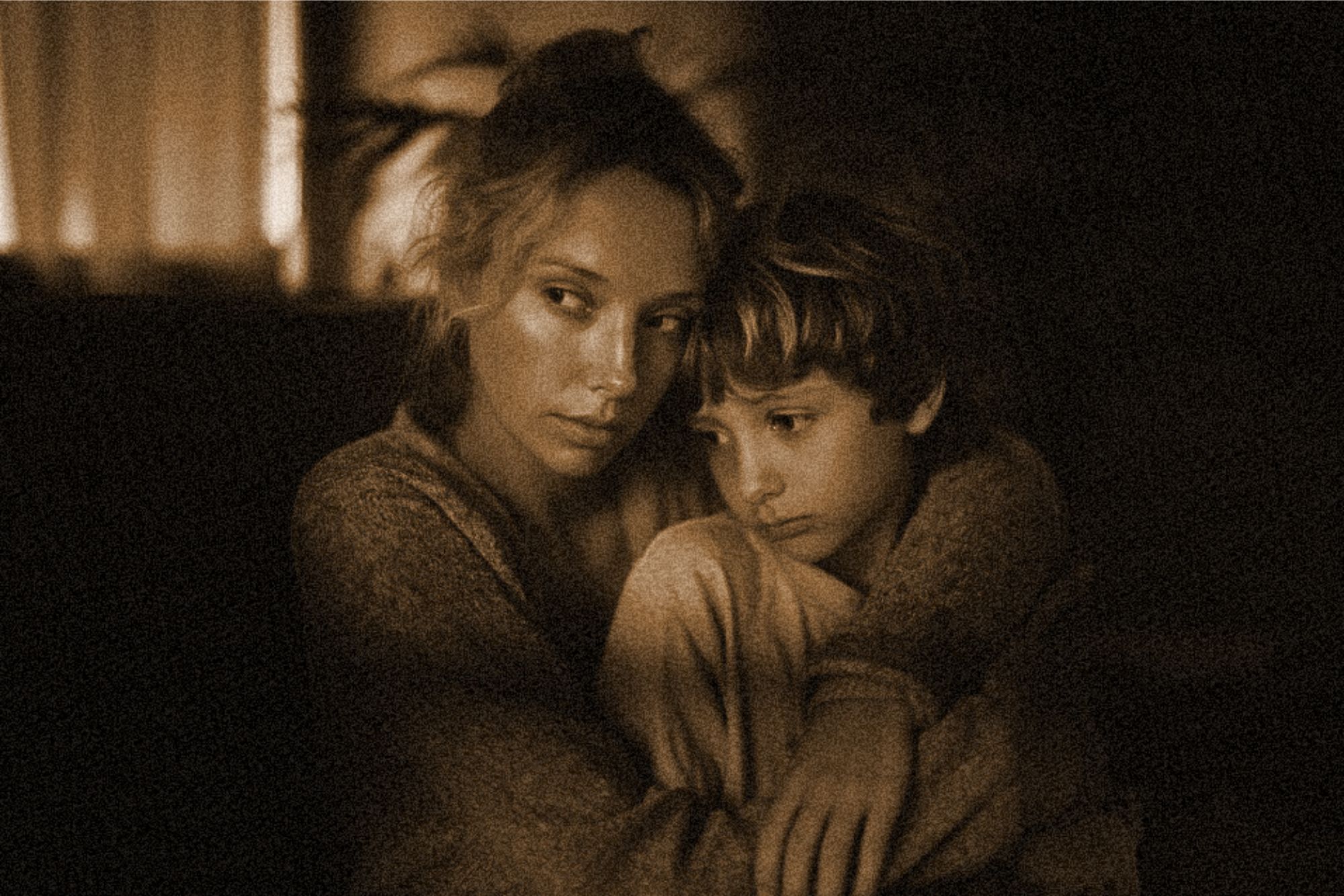 A woman hugs a young boy closely, both looking somber and thoughtful. The warm, sepia-toned lighting and soft focus create an intimate, comforting atmosphere.
