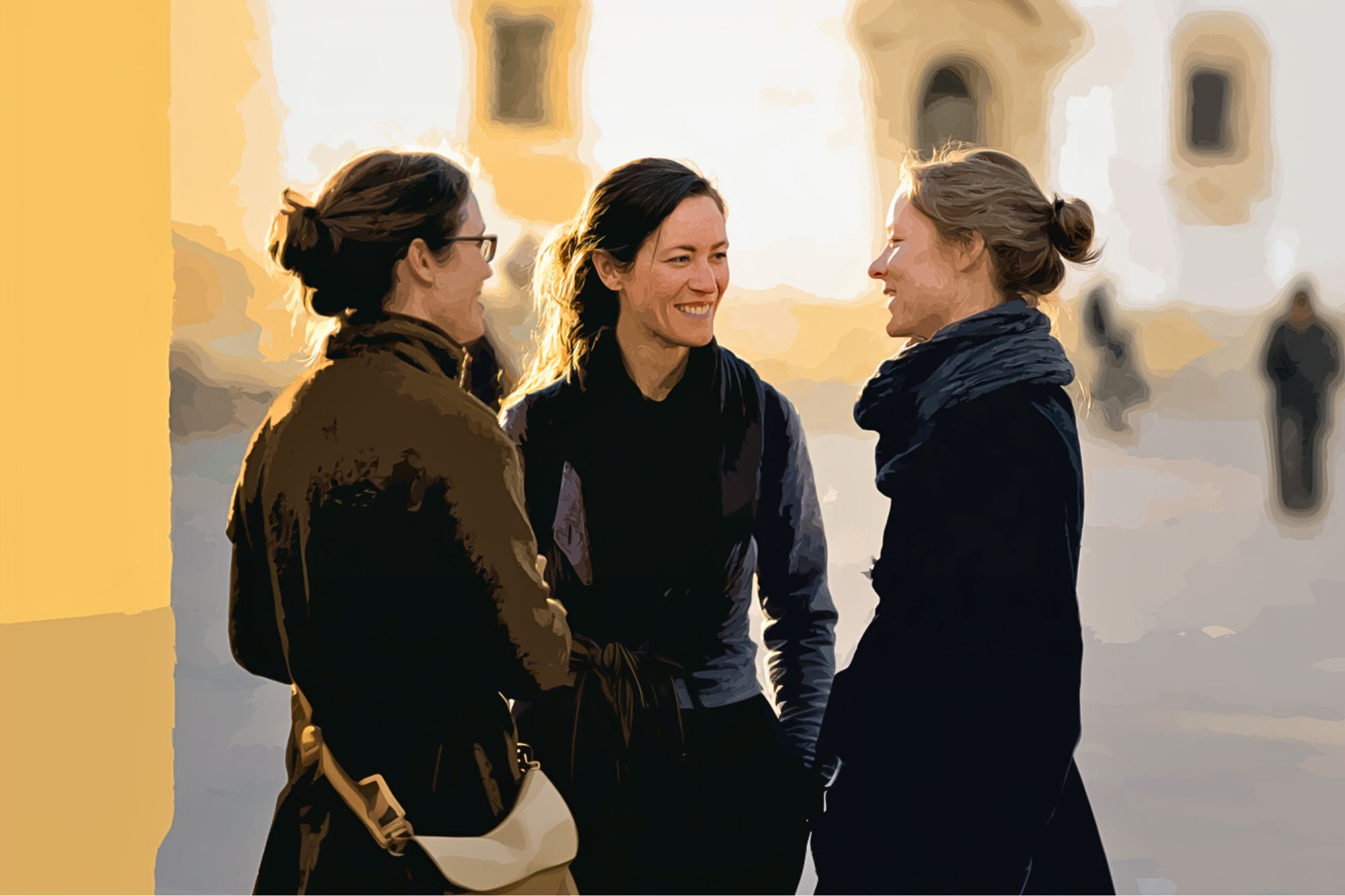Three women stand outdoors in conversation, smiling at each other. They wear dark coats and have their hair tied back. The background is softly blurred, suggesting a bright, sunlit day in an urban setting.