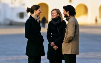 Three people wearing coats stand outside in conversation, smiling at each other. The background is softly blurred, with white and yellow buildings and arched doorways visible in the distance.