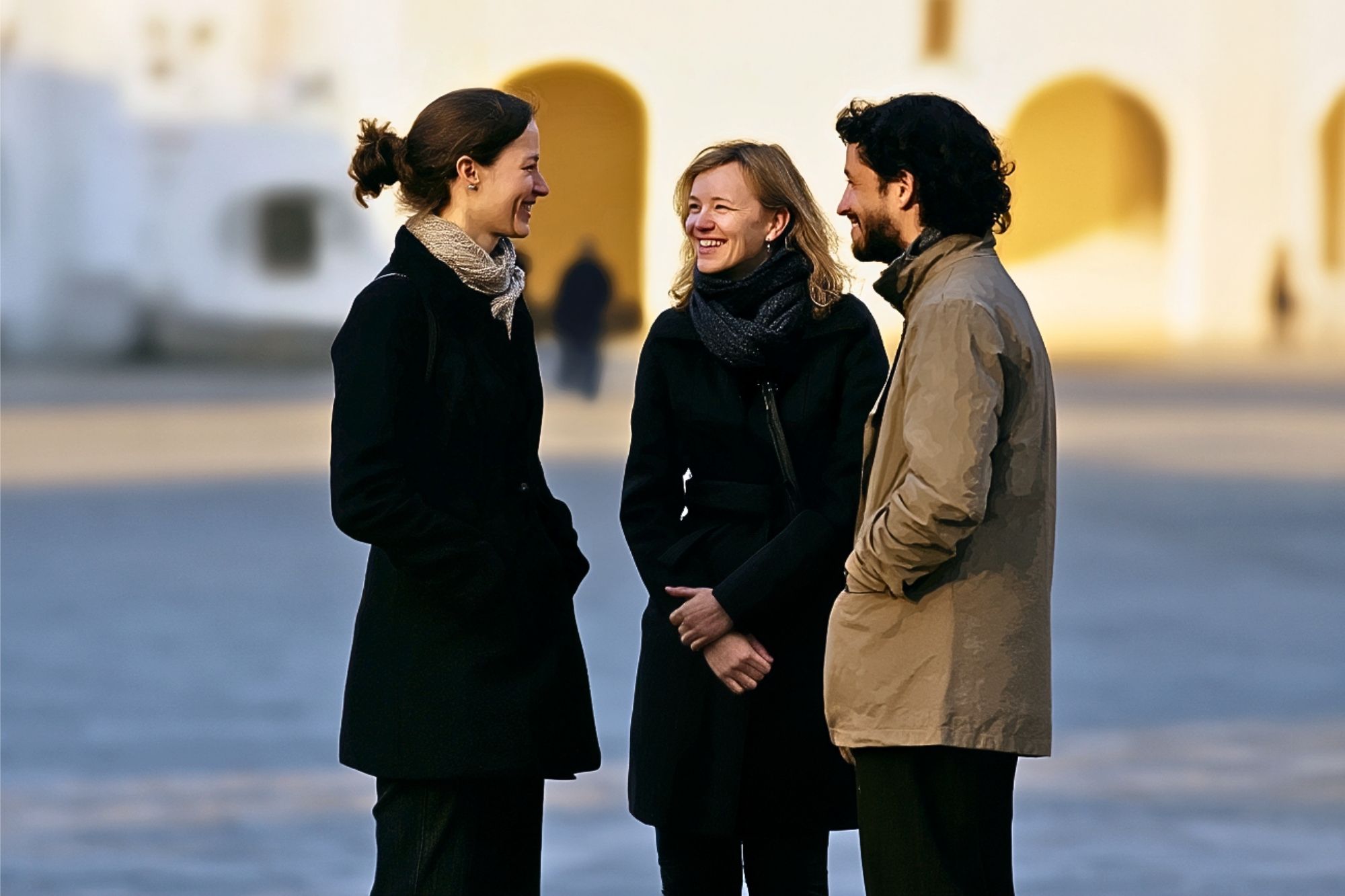 Three people wearing coats stand outside in conversation, smiling at each other. The background is softly blurred, with white and yellow buildings and arched doorways visible in the distance.