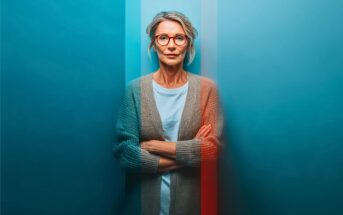 A middle-aged woman with glasses and gray hair stands against a blue wall with her arms crossed, wearing a gray cardigan and white shirt. Vertical blue and red light streaks overlay the image.
