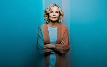 A woman with blonde hair stands with arms crossed, wearing a light shirt and cardigan. Blue and orange lighting creates a dramatic split-color effect on her and the background.