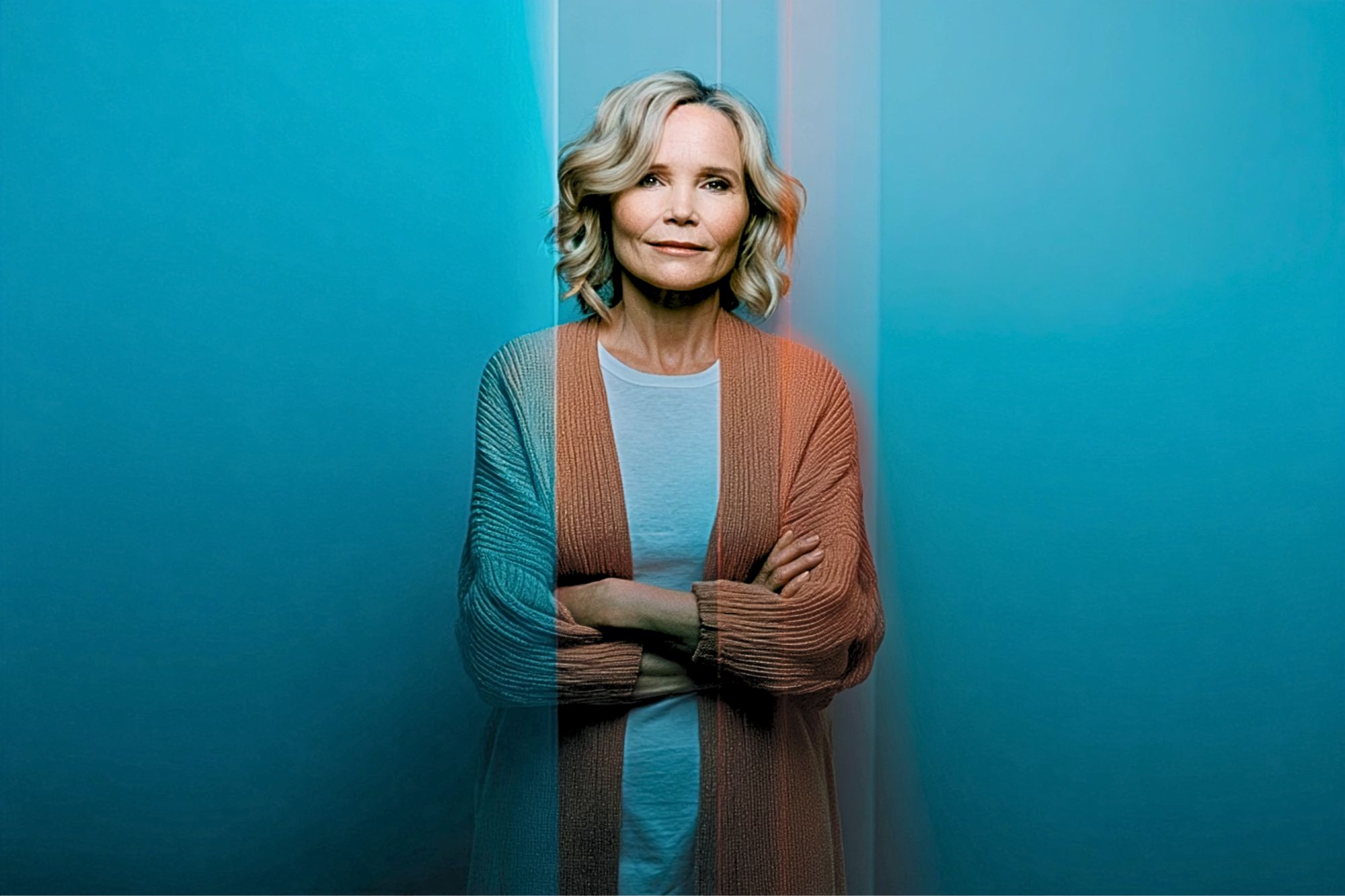 A woman with blonde hair stands with arms crossed, wearing a light shirt and cardigan. Blue and orange lighting creates a dramatic split-color effect on her and the background.