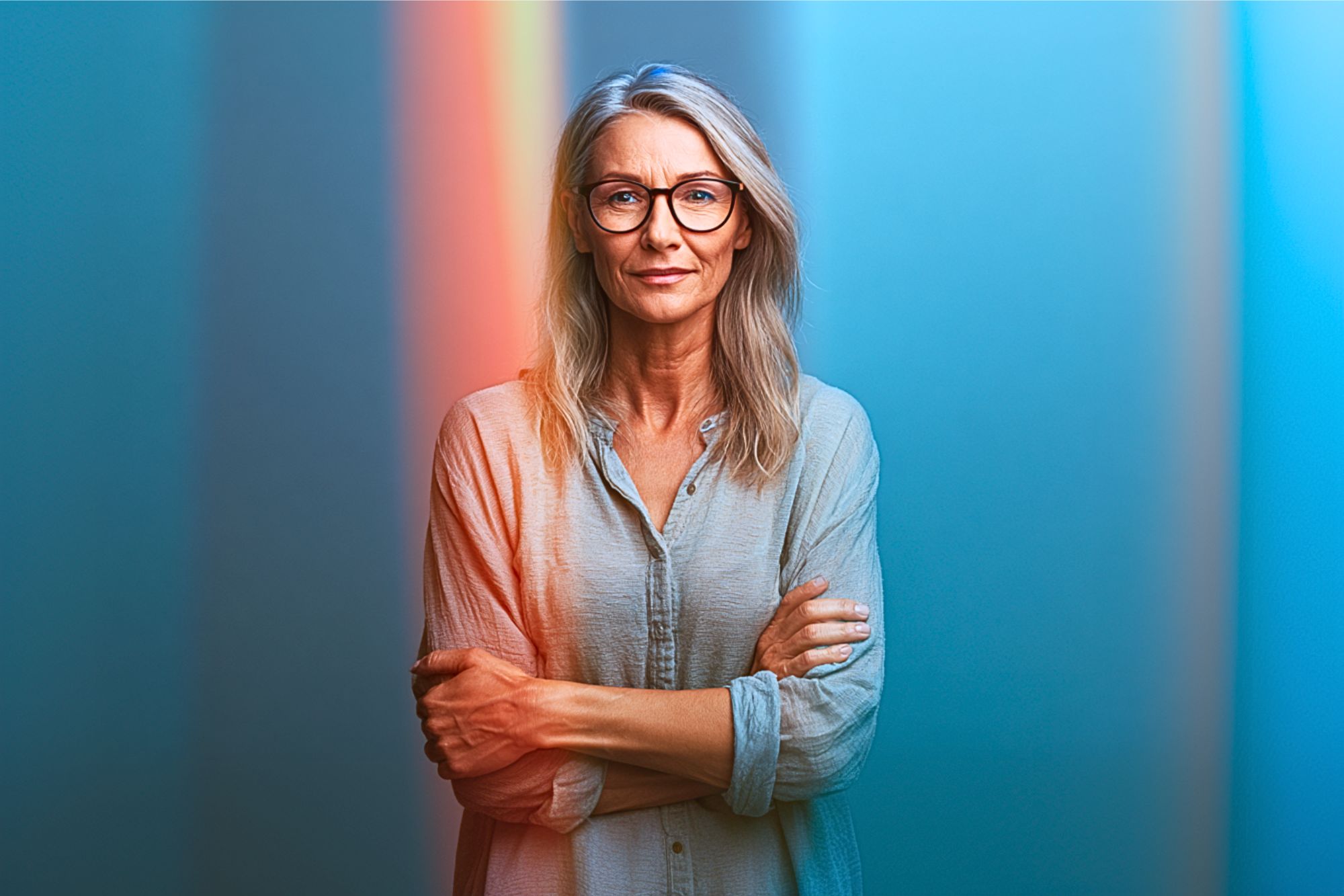 A confident middle-aged woman with long blond hair and glasses stands with arms crossed, wearing a light blue shirt, against a blue background with vertical colored light streaks.