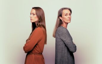 A young woman and an older woman stand back-to-back with arms crossed, both looking away from each other with serious expressions against a light, neutral background.