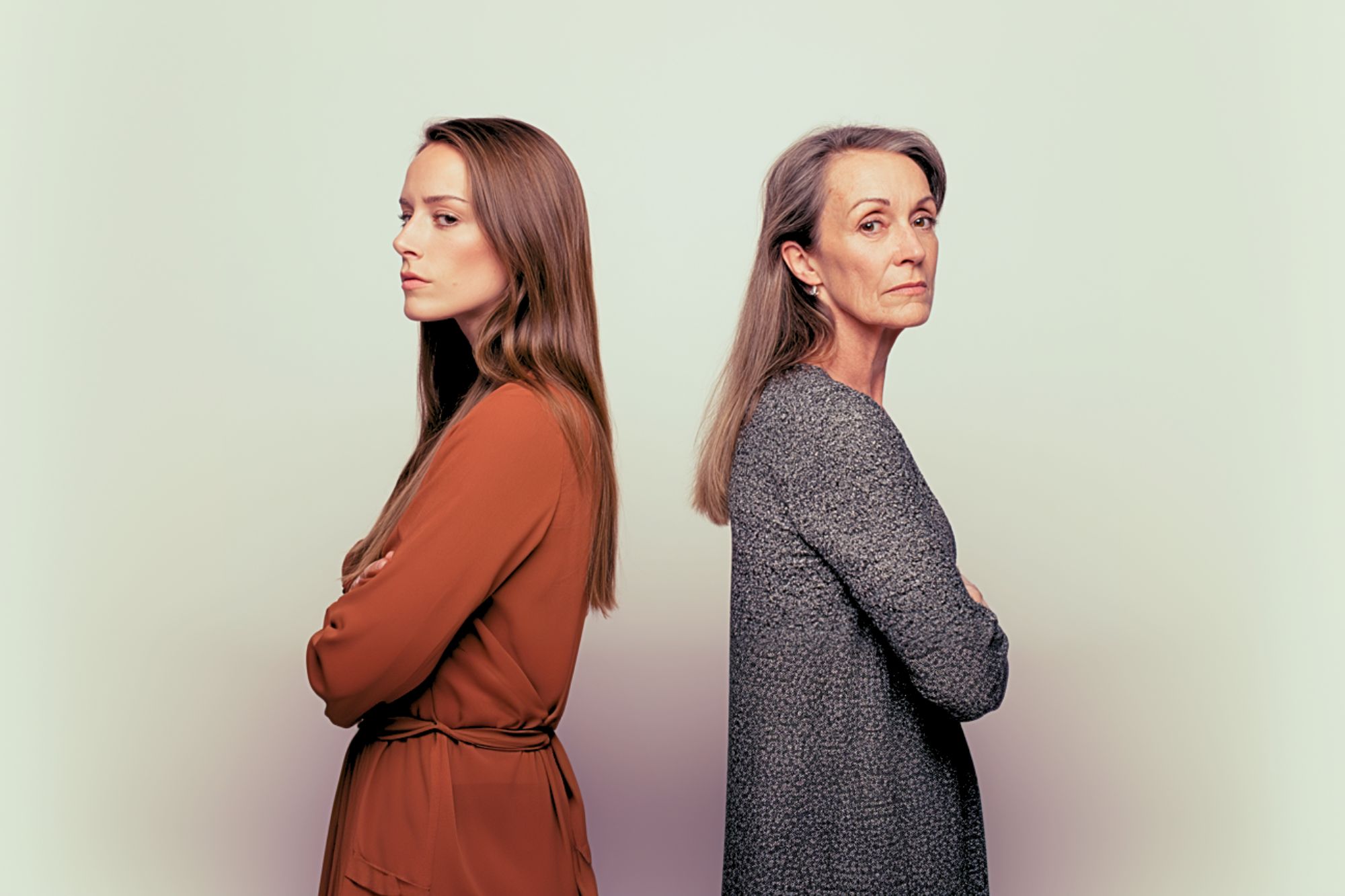 A young woman and an older woman stand back-to-back with arms crossed, both looking away from each other with serious expressions against a light, neutral background.