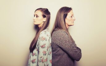 Two women stand back to back, looking away from each other with serious expressions. One is younger, wearing a floral top, and the other is older, wearing a knit sweater. The background is plain and beige.