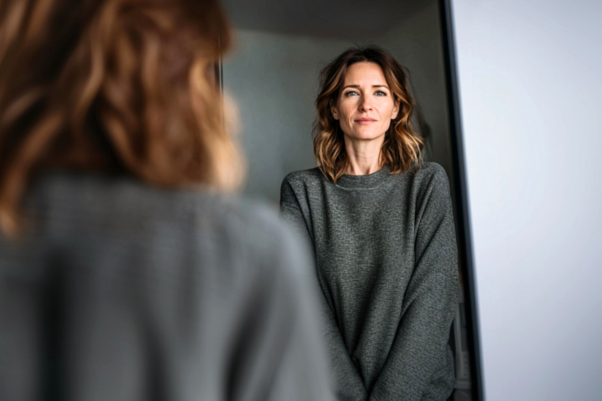 A woman with shoulder-length wavy hair and a gray sweater looks at her reflection in a mirror, appearing calm and thoughtful in a softly lit room.