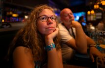 A woman with curly hair and glasses rests her chin on her hand, looking thoughtful, while a man sits beside her in a dimly lit restaurant or bar.
