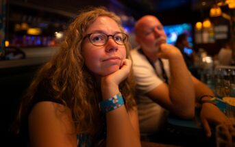 A woman with curly hair and glasses rests her chin on her hand, looking thoughtful, while a man sits beside her in a dimly lit restaurant or bar.