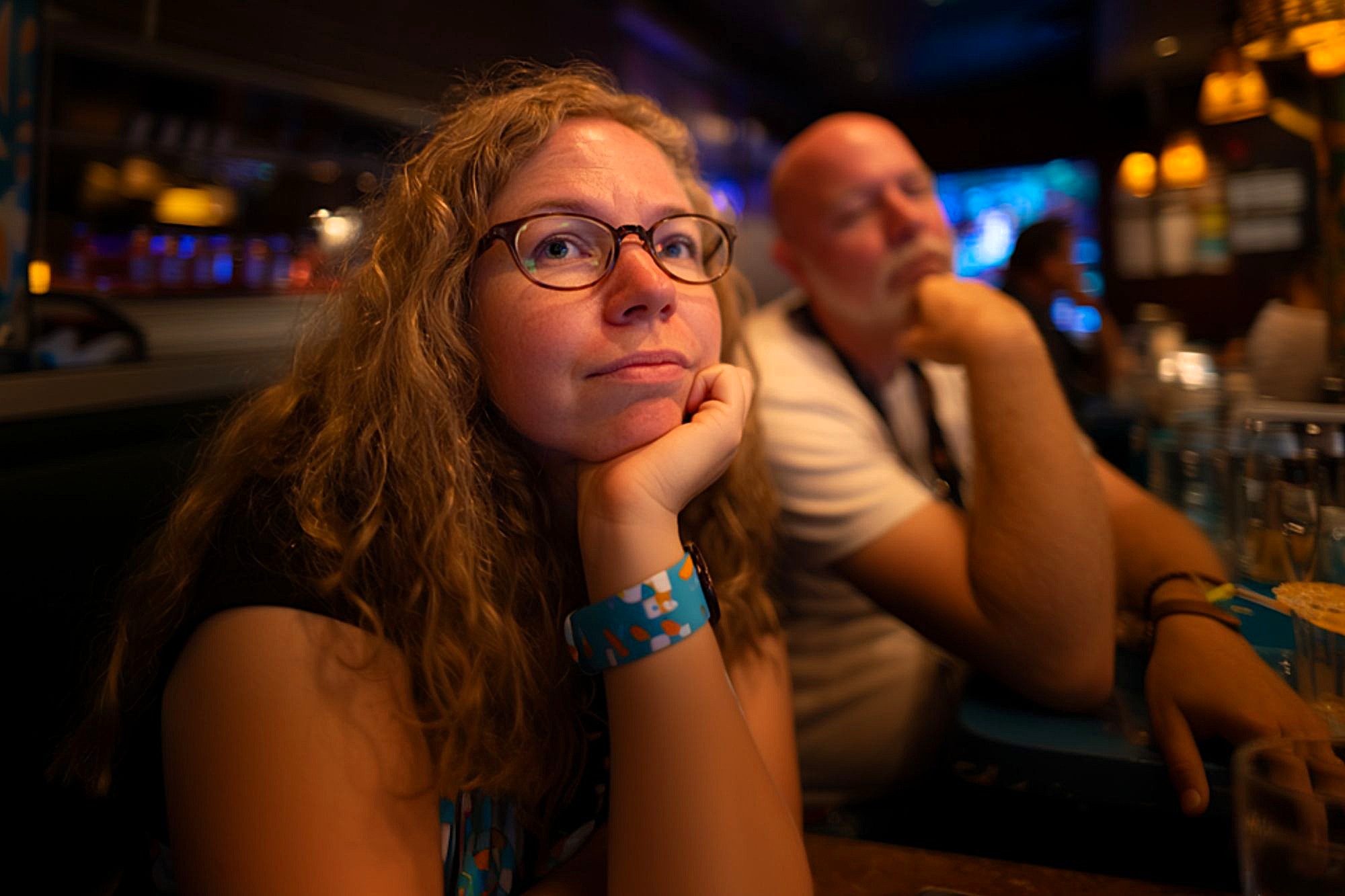 A woman with curly hair and glasses rests her chin on her hand, looking thoughtful, while a man sits beside her in a dimly lit restaurant or bar.