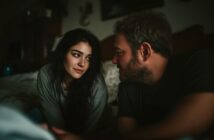 A woman with long dark hair and a man with light hair are sitting closely together indoors, gazing into each other's eyes with soft expressions in a dimly lit room.