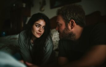 A woman with long dark hair and a man with light hair are sitting closely together indoors, gazing into each other's eyes with soft expressions in a dimly lit room.