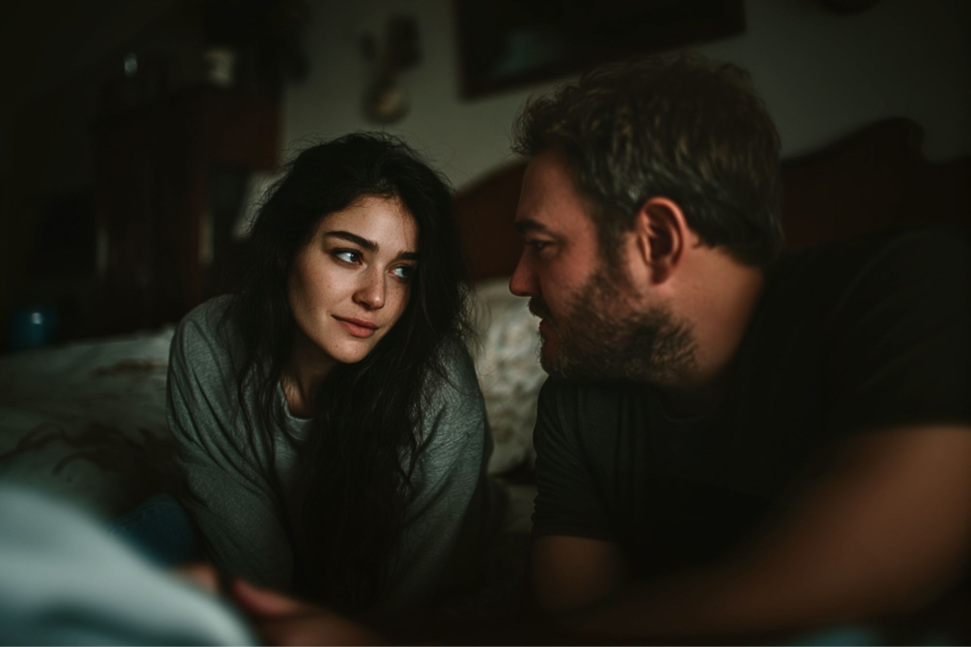 A woman with long dark hair and a man with light hair are sitting closely together indoors, gazing into each other's eyes with soft expressions in a dimly lit room.