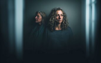 Two women stand close together in a dimly lit, shadowy room, both wearing dark sweaters. One woman looks to the side with a neutral expression; the other faces forward, gazing thoughtfully ahead.