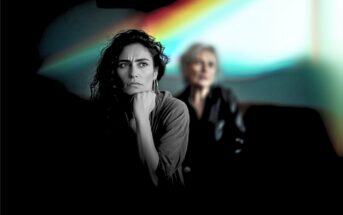 A woman with curly hair looks pensive in the foreground, while an older woman sits blurred in the background. A colorful light streak is visible behind them.