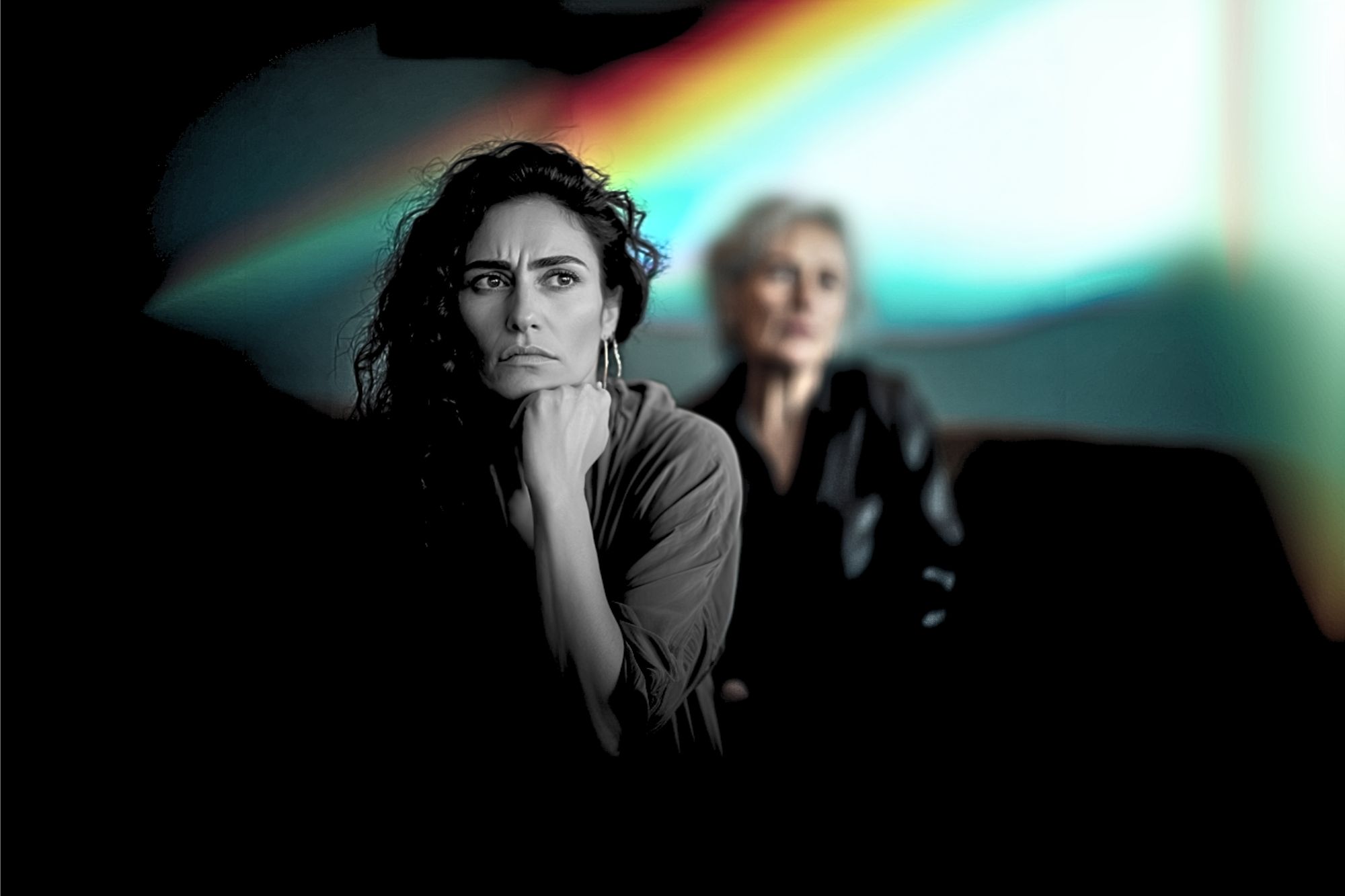 A woman with curly hair looks pensive in the foreground, while an older woman sits blurred in the background. A colorful light streak is visible behind them.