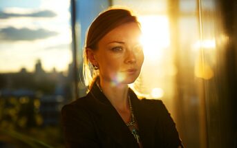 A woman with light hair stands indoors by a window at sunset, sunlight shining on her face and casting a warm glow. She looks thoughtful, with arms crossed, and city buildings are blurred in the background.