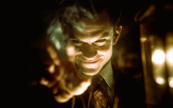 A man with a sinister smile is dramatically lit from below, reaching forward with an outstretched hand. His face is partially shadowed, creating a mysterious and intense atmosphere. Warm light glows to the right.